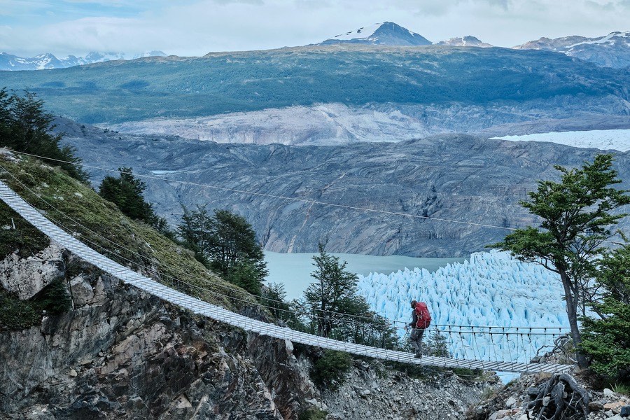 A hiker crossing a suspension bridge above a glacier and its turquoise meltwater lake
